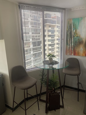 A room with a glass table and two chairs, a window with blinds, a potted plant, and a painting on the wall.