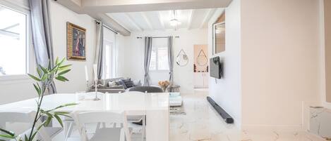 A modern living room with a white sofa, dining table, and chairs, a TV mounted on the wall, and a kitchen in the background.