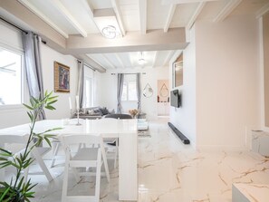 A modern living room with a white sofa, dining table, and chairs, a TV mounted on the wall, and a kitchen in the background. - BEAUSOLEIL FAMILY, WEDDING & EVENT VENUE IN FRANCE (Beausoleil)