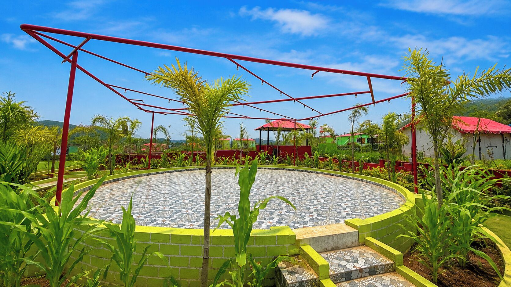 A circular outdoor area with a tiled floor, surrounded by greenery and a red metal structure.