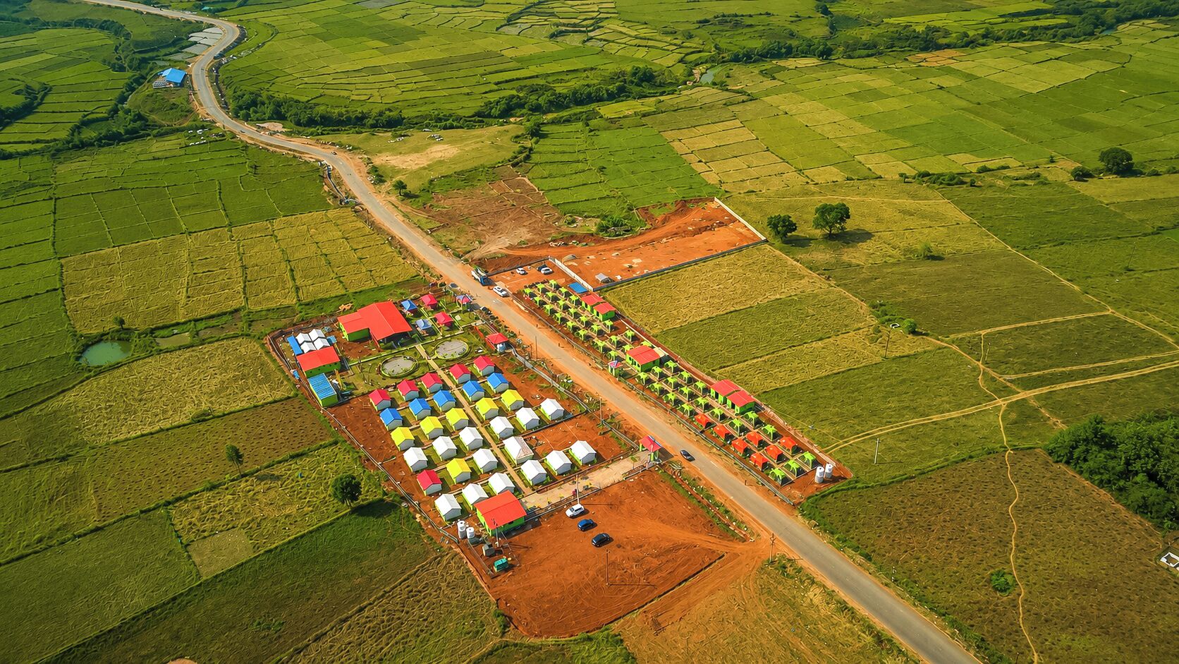 An aerial view of a farm with multiple green fields, a road, and a large building with colorful roofs.