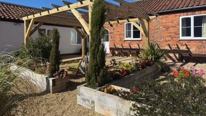 Une maison en briques avec une pergola en bois, un jardin où poussent différentes plantes et fleurs, et un ciel bleu dégagé.