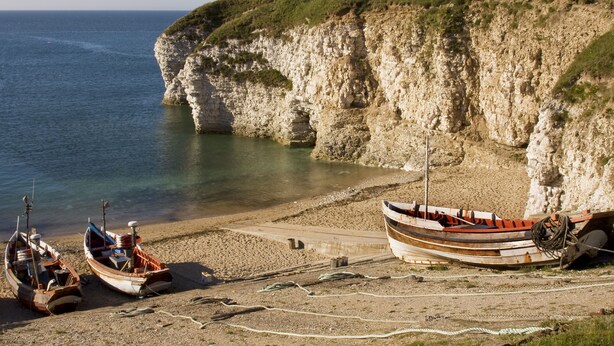 Un paysage côtier avec des bateaux sur une plage de sable, des falaises en arrière-plan et une eau calme.