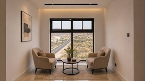 A room with two chairs facing a large window, a small table with a vase, and a framed picture on the wall.