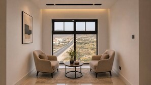 Una habitación con dos sillas orientadas a un ventanal grande, una mesita con un florero y un cuadro en la pared.