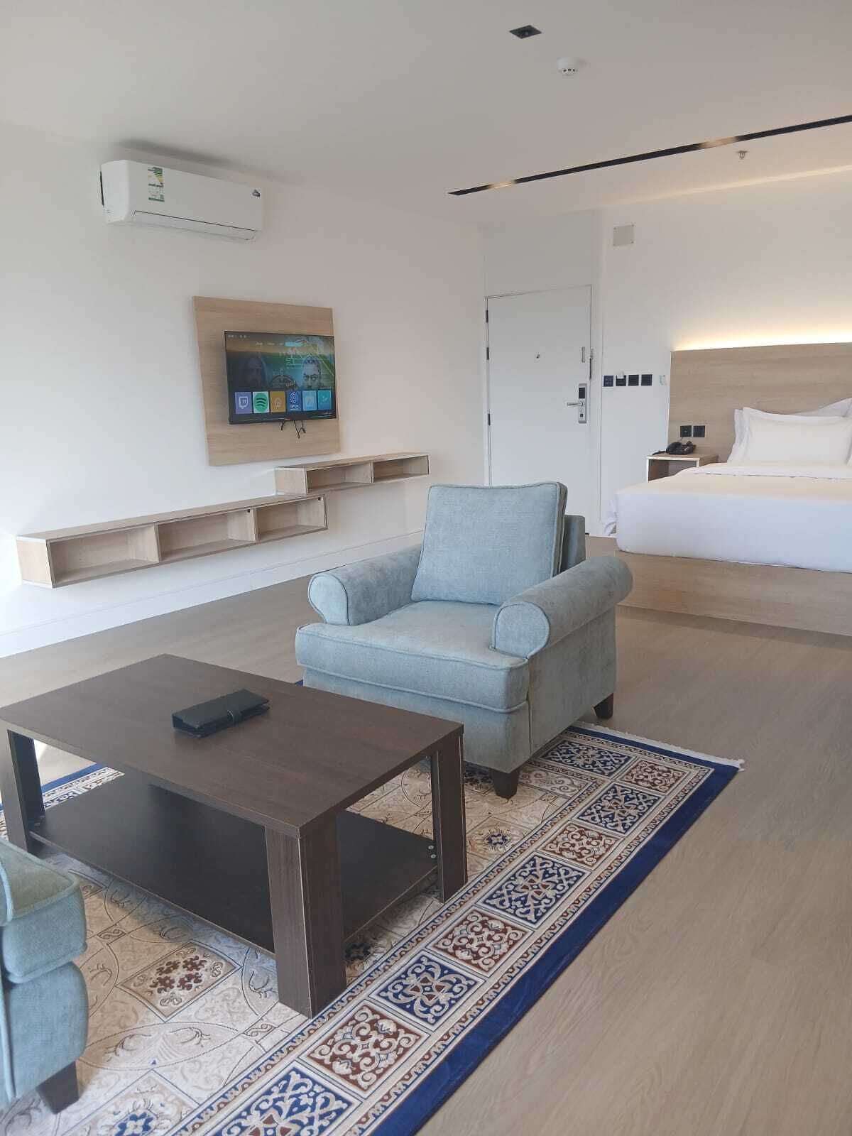 Una habitación de hotel moderna con sofá, una mesita de centro, televisor montado en la pared, cama y puerta.