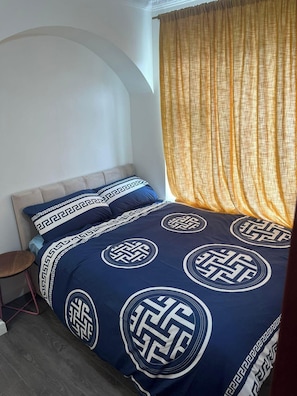 Un dormitorio con cama, mesita de noche, silla y ventana con cortinas.
