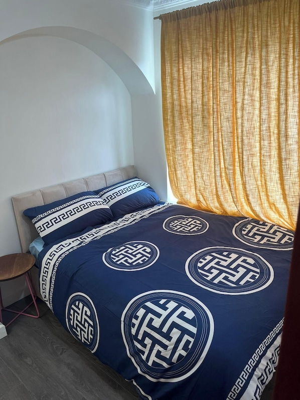 Un dormitorio con cama, mesita de noche, silla y ventana con cortinas.
