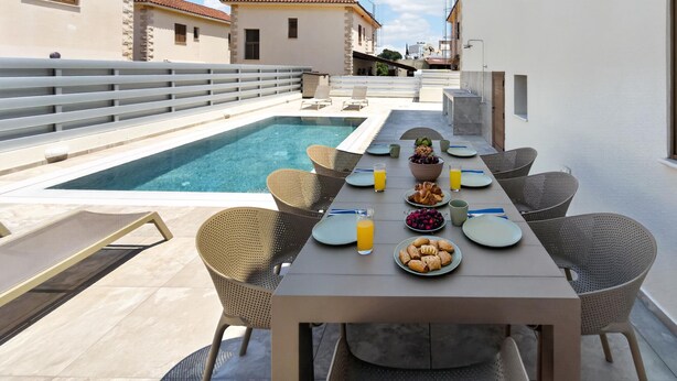 A poolside dining area with a table set for two, surrounded by wicker chairs, a pool, and a white building with a tiled roof in the background. - Delina Coastal Villa Steps from Everything (Protaras)