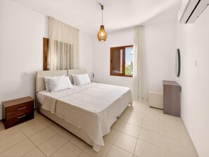 A bedroom with a bed, a nightstand, a bedside table, a window with curtains, and a wall-mounted air conditioning unit. - Delina Coastal Villa Steps from Everything (Protaras)