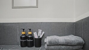 Casa de banho com azulejos cinzentos, uma pia, duas garrafas de shampoo e condicionador, uma toalha e um porta-escova de dentes.