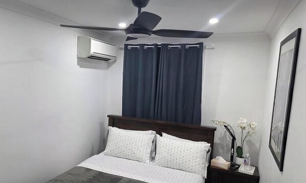 Une chambre à coucher avec un lit, un ventilateur de plafond, une fenêtre avec des rideaux, une table de chevet avec une lampe et un tableau encadré accroché au mur.