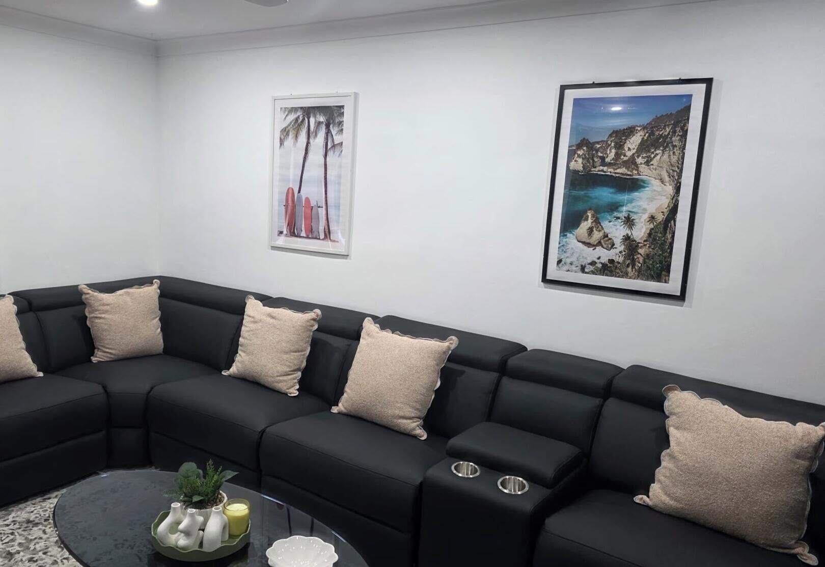 Ein schwarzes Ecksofa mit beigen Kissen, ein Glastisch mit Dekoration und zwei gerahmte Landschaftsbilder an der Wand.