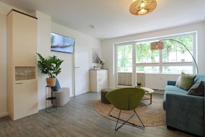 Ein modernes Wohnzimmer mit Sofa, grünem Sessel, Couchtisch, Fernseher und großem Fenster.