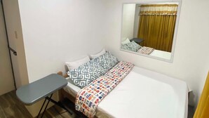 Ein Einzelbett mit gemusterten Kissen, ein Nachttisch, ein Spiegel und ein Fenster mit Vorhängen.