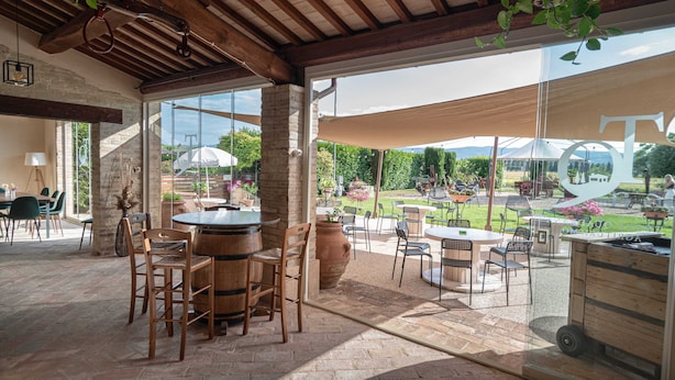 Un patio exterior con mesas y sillas, un barril de madera y una pared de vidrio que ofrece vistas del jardín.