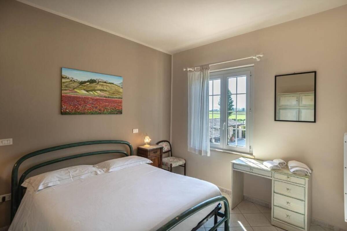 A bedroom with a bed, a desk, a chair, a window with curtains, a painting on the wall, and a mirror.