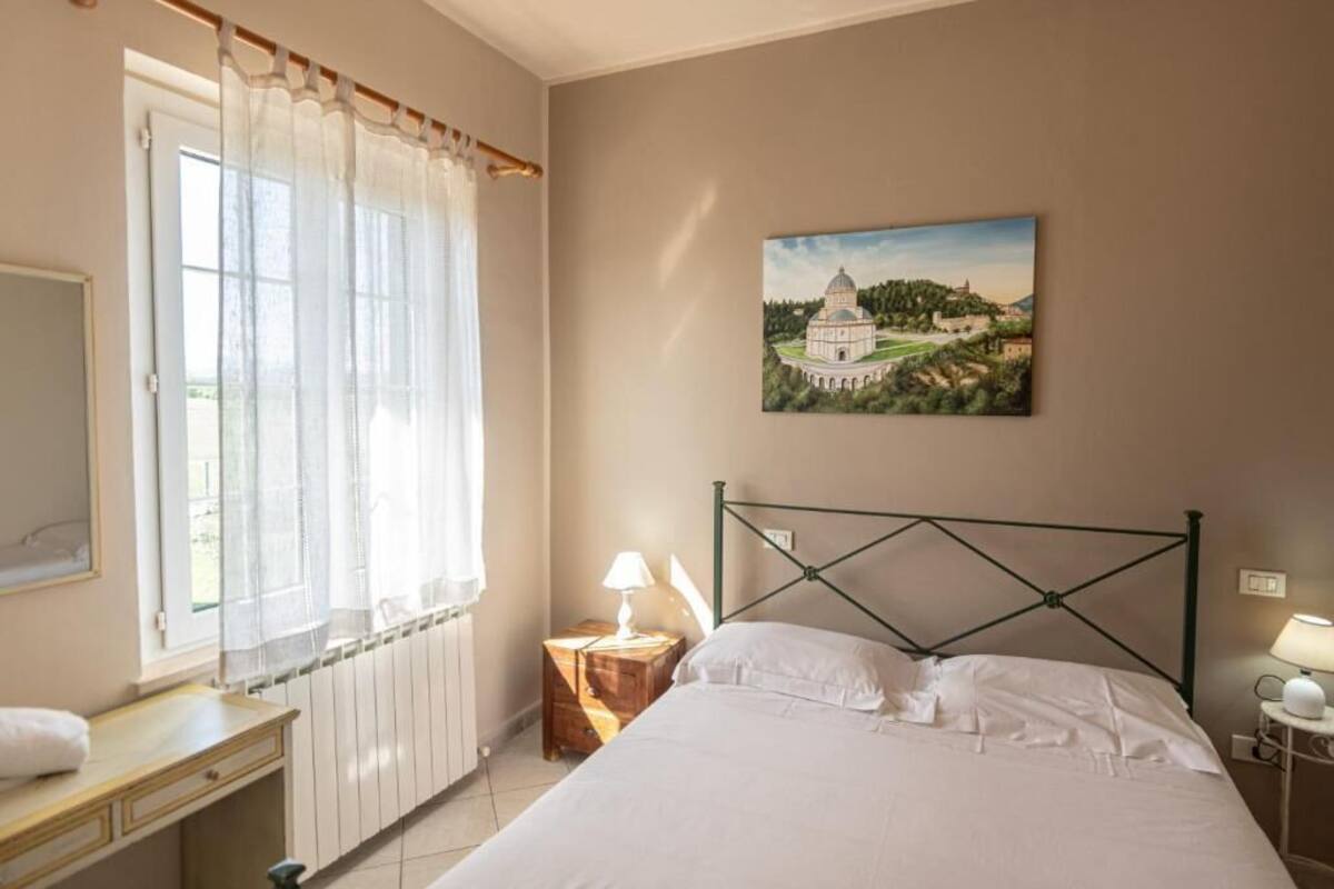 A bedroom with a bed, a nightstand, a lamp, a window with curtains, a radiator, and a painting on the wall.
