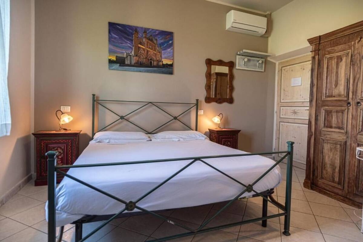 A room with a bed, two bedside tables, a wall-mounted air conditioner, a wooden wardrobe, and a painting of a building.