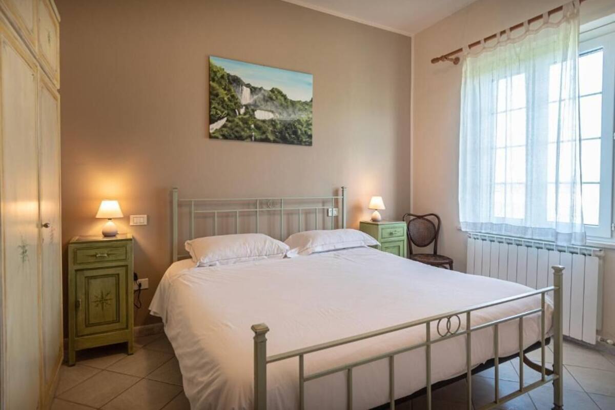 A bedroom with a single bed, two bedside tables, a nightstand, a chair, a window with curtains, and a painting on the wall.