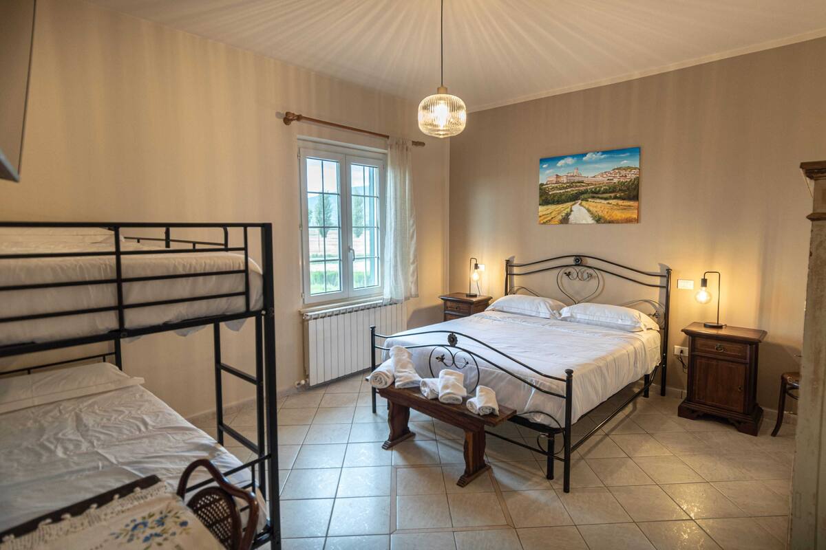 A bedroom with a bunk bed, a single bed, a bedside table with a lamp, a window with curtains, and a painting on the wall.