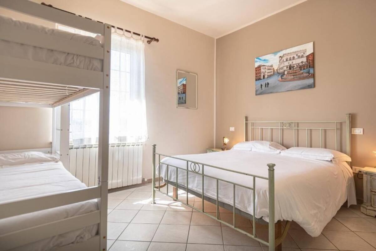 A bedroom with bunk beds, a single bed, a radiator, a window with curtains, a wall-mounted picture, and bedside lamps.