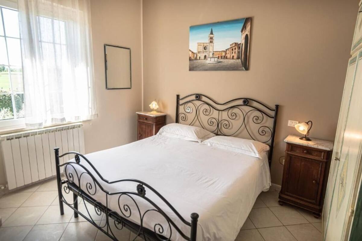 A bedroom with a bed, a window with curtains, a wall-mounted picture, bedside tables with lamps, and a radiator.