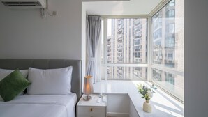 Habitación de hotel con cama, mesita de noche, lámpara y ventana con vista a edificios altos.