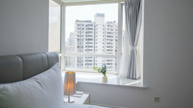 Una habitación de hotel moderna con un ventanal grande, un sofá gris, una mesita de noche con una lámpara y un pequeño jarrón con flores.
