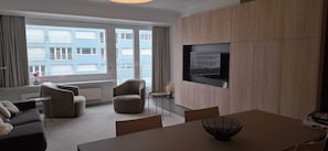 Ein modernes Wohnzimmer mit Sofa, Sesseln, einem Esstisch, einem an der Wand befestigten Fernseher und großen Fenstern mit Blick auf ein Gebäude.