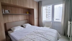 Ein Schlafzimmer mit Bett, Stuhl, einer Kommode mit Gegenständen und einem Fenster mit Blick auf Gebäude.