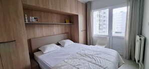 Ein Schlafzimmer mit Bett, Stuhl, einer Kommode mit Gegenständen und einem Fenster mit Blick auf Gebäude.