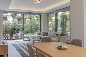 A modern living room with a sofa, a dining table, and a view of the outdoors through large windows. - Private Pool Villa | Walk to Beach & Voula Square (Voula)
