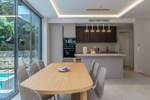 A modern kitchen with a central island, a dining area with a table and chairs, and a large sliding glass door leading to a pool. - Private Pool Villa | Walk to Beach & Voula Square (Voula)