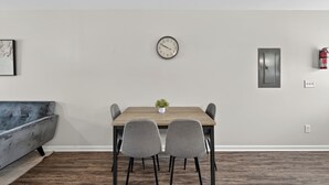 A dining area with a wooden table, two grey chairs, a small potted plant, a wall clock, a fire extinguisher, and a grey sofa. - Cozy 2br Stay Near Work & Dining (Clarksville)