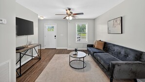 A living room with a ceiling fan, a flat-screen TV, a dark sofa, a round coffee table, and a window with blinds. - Cozy 2br Stay Near Work & Dining (Clarksville)
