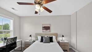 A bedroom with a bed, desk, chair, bedside tables, a ceiling fan, and a window with blinds. - Cozy 2br Stay Near Work & Dining (Clarksville)