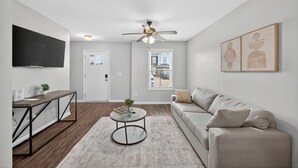 A modern living room with a flat-screen TV, a ceiling fan, a sofa, a coffee table, and a view of the city through a window. - Bright, Modern 2 bedroom Home Away From Home (Clarksville)