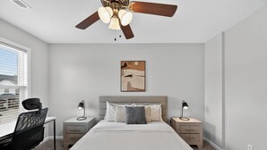 A bedroom with a bed, desk, chair, nightstands, lamps, a ceiling fan, and a painting on the wall. - Bright, Modern 2 bedroom Home Away From Home (Clarksville)