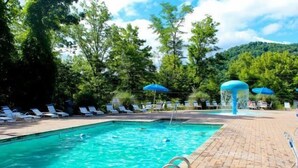 A swimming pool with lounge chairs and umbrellas, surrounded by trees and a hill in the background. - Pool table, hot tub, indoor sauna, outdoor spaces, big kitchen in Gatlinburg! (Gatlinburg)