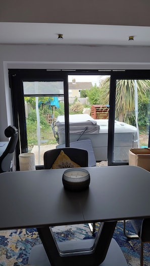 Una habitación con una mesa, una silla, un ventanal y vista a un patio trasero con jacuzzi y vegetación.