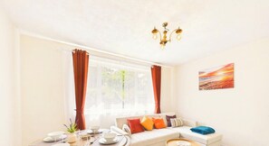 Ein Zimmer mit einer Couch, einem Esstisch gedeckt mit Tellern und Tassen, einem Fenster mit Vorhängen und einem Bild an der Wand.