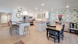 A modern kitchen with white cabinetry, a central island, and a dining area with a wooden table and chairs. - Sea Oats 117 | 2BR/2BA Boca Grande Gulf Front Luxe (Boca Grande)