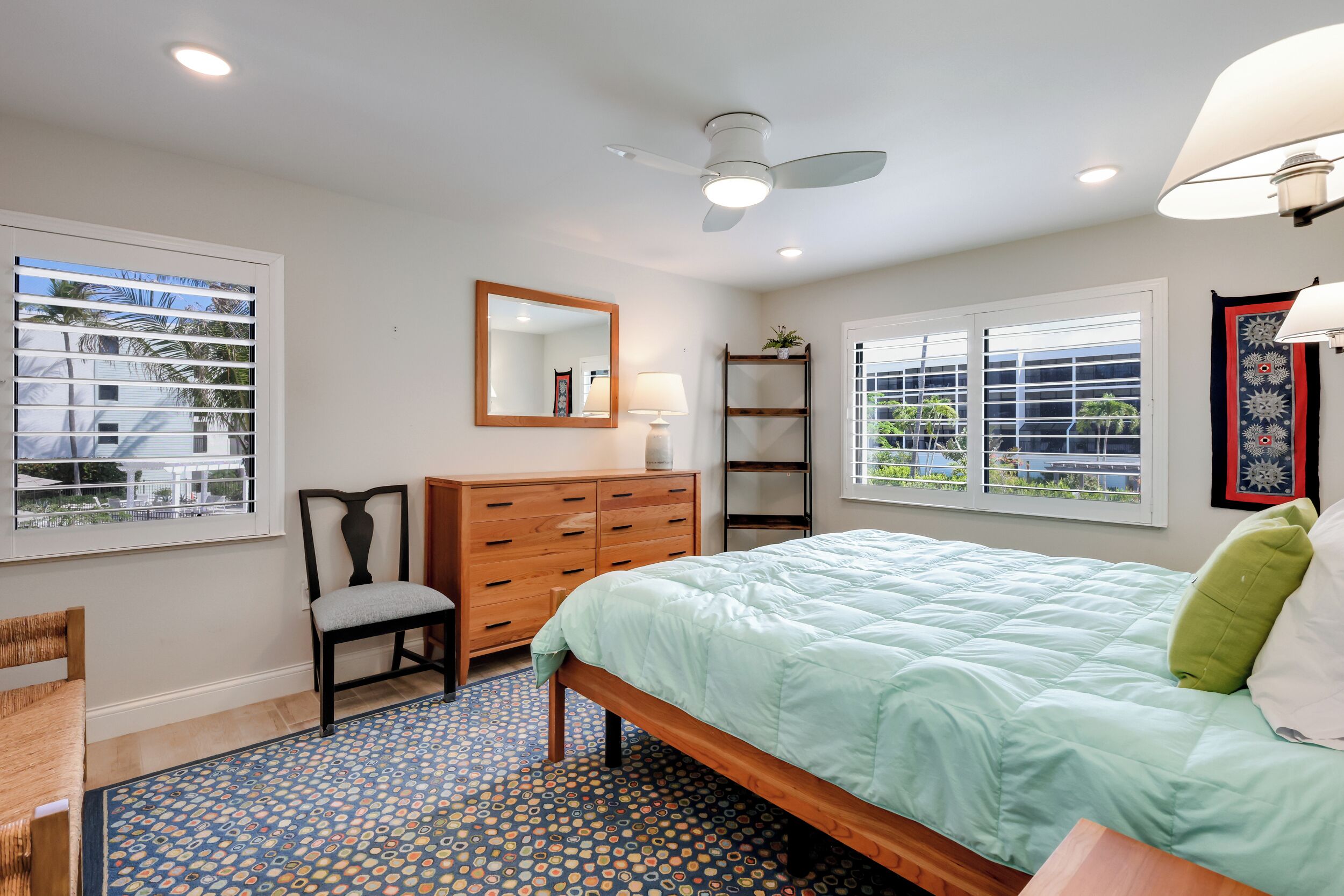 A bedroom with a bed, a chair, a dresser, a mirror, and two windows with blinds.