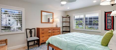 A bedroom with a bed, a chair, a dresser, a mirror, and two windows with blinds.