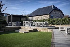 A backyard with a stone building, outdoor grill, and a grassy area with steps leading to a patio.