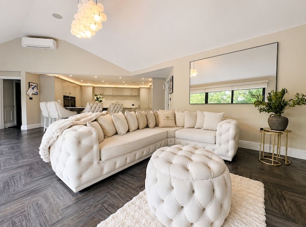 A spacious living room with a large sectional sofa, a round ottoman, a chandelier, and a large mirror.