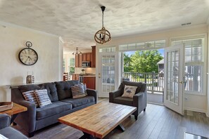 A spacious living area with a sofa, armchair, and coffee table, a clock on the wall, and a view to the outside.