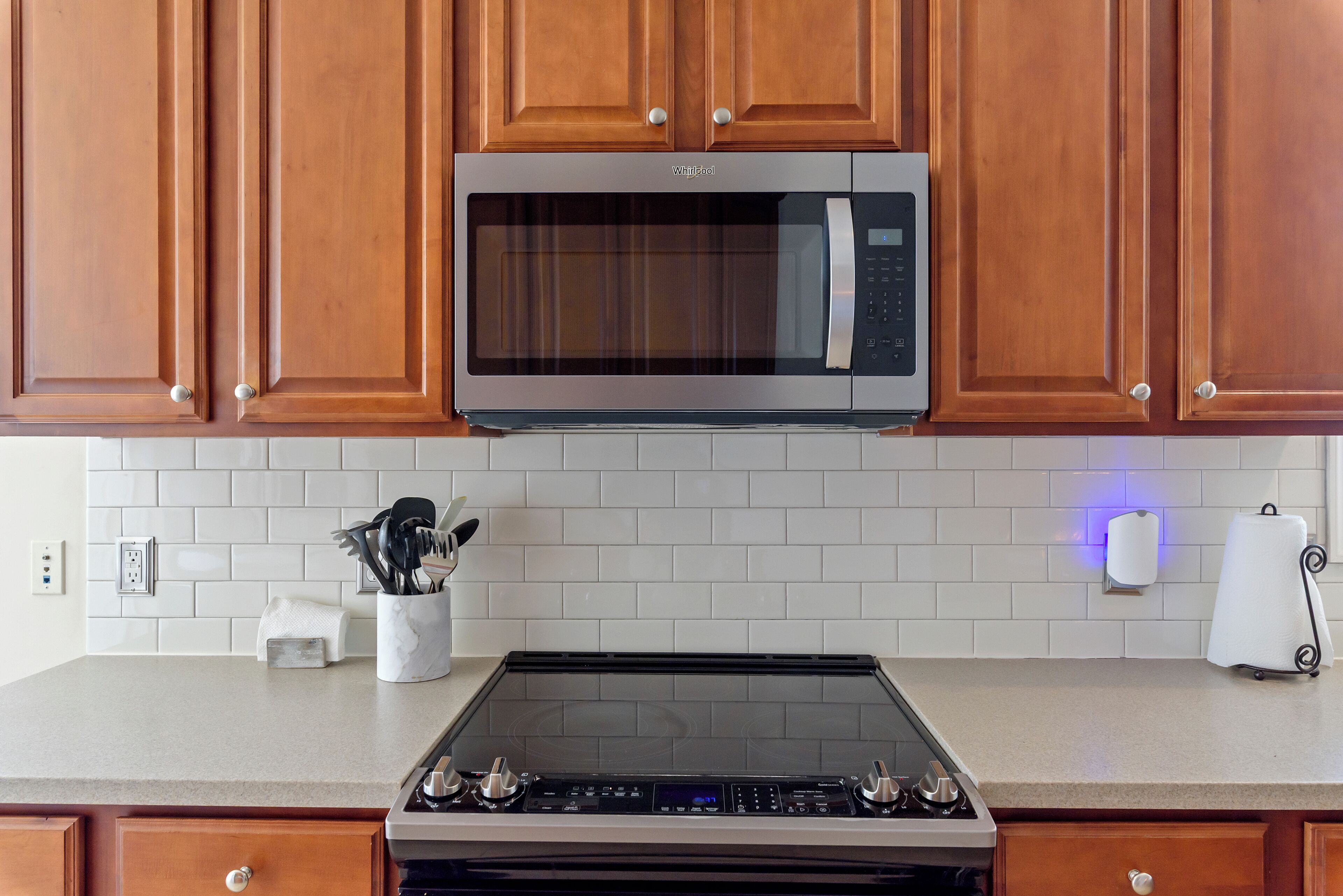 Une cuisine avec des meubles en bois, un four à micro-ondes en acier inoxydable, un four encastré, un crédence en carrelage blanc et un plan de travail doté d’un distributeur d’essuie-tout et d’un porte-couteaux.