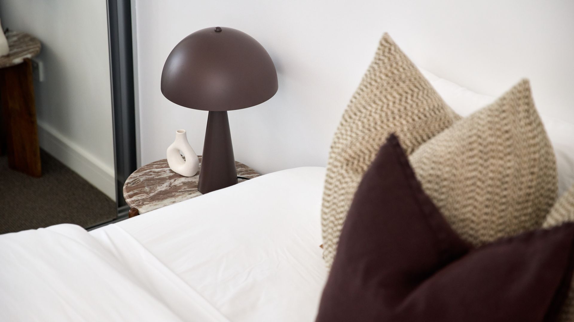 A neatly made bed with white linens, a dark brown lamp on a round side table, and a vase with a white, abstract design.
